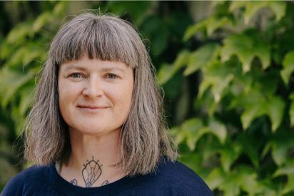 Porträtfoto weibliche Person mit halblangen Haaren und blauem Oberteil, iim Hintergrund grüne Pflanzen