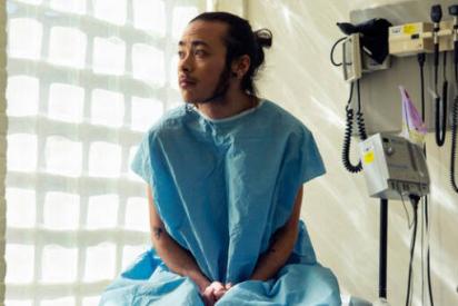 A genderqueer person sitting in a hospital gown sitting in an exam room