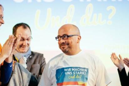 Drei Personen applaudieren einer Person im weißen T-Shirt mit der Aufschrift &quot;Wir machen uns stark! Und du?&quot;