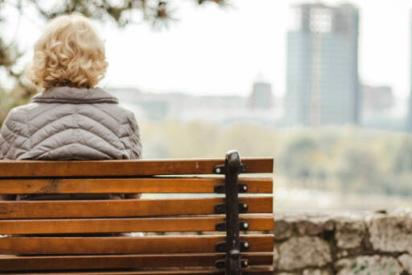 Altere Frau von hinten, sie sitzt auf einer Bank und blickt in die Ferne.